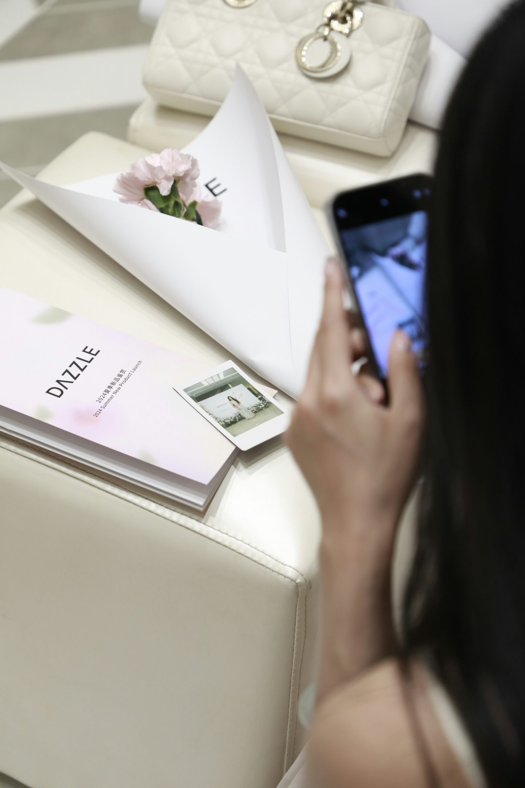 Blick über die Schulter einer dunkelhaarigen Frau, die einige Objekte, die vor ihr liegen, mit der Handykamera fotografiert. Der Fokus des Bilds liegt auf den Objekten. Es liegen eine weiße Clutch, eine in Papier eingepackte Blume mit zartrosa Blüten, ein zartrosa Leporello der Marke Dazzle und ein Polaroid nebeneinander auf einem weißen Ledersitzwürfel.