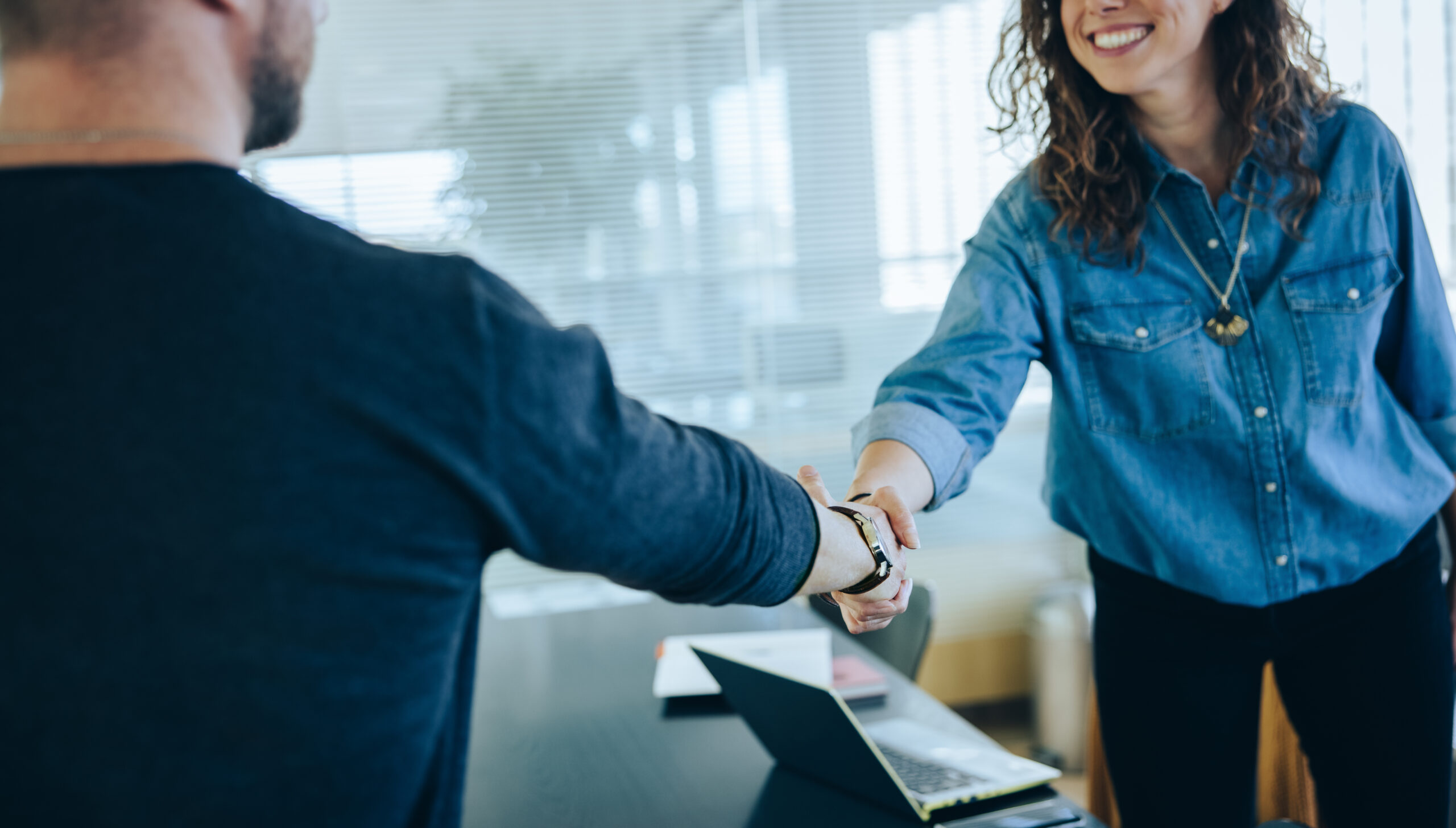 Eine Frau und ein Mann schütteln Hände in einer Arbeitsumgebung mit aufgeklapptem Laptop im Hintergrund.